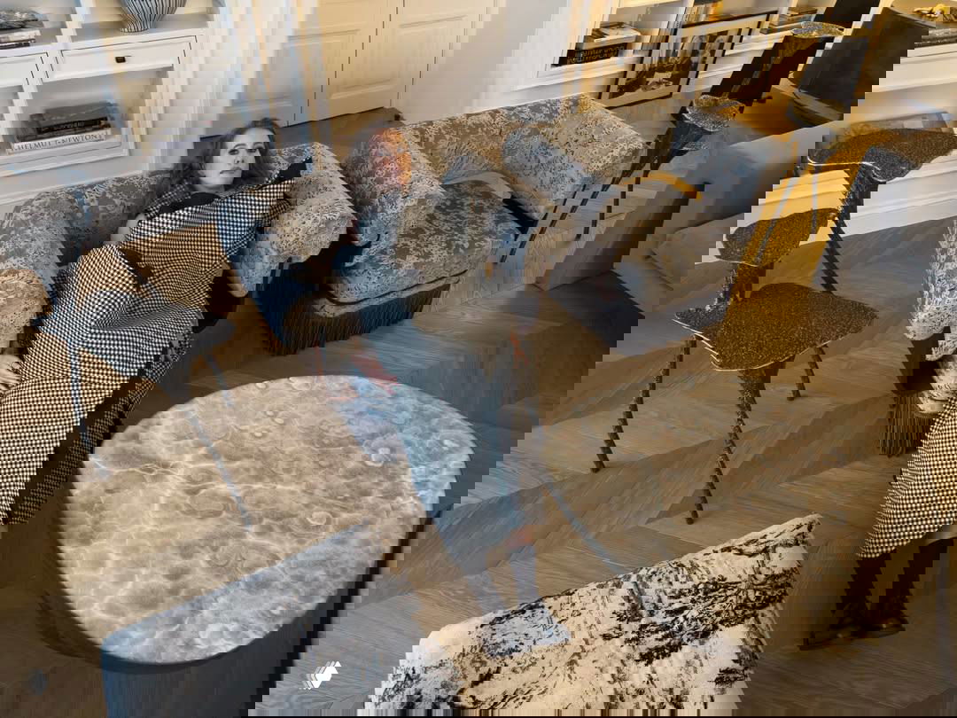 Интерьер с фрагментом паркета «ёлочка», низкими креслами и круглым мраморным столом; в кадре расположена женщина на кресле в клетчатом наряде