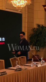 Бурак Озчивит в тёмном костюме у стола на пресс‑конференции в Москве; видна люстра, микрофоны и ватермарк Woman.ru на фото.