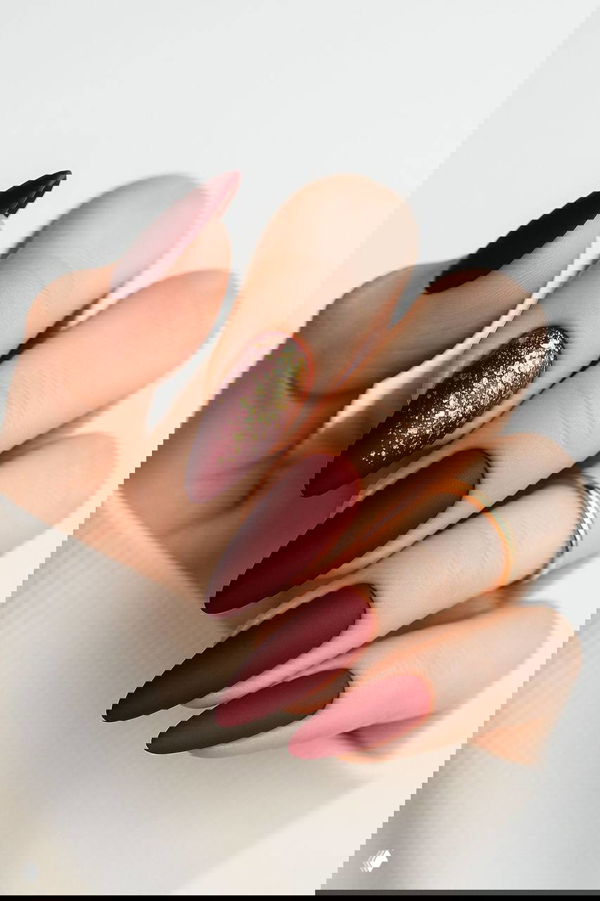 Матовый бордово-красный удлинённый маникюр с золотыми блёстками на одном ногте и элегантными кольцами на пальцах — трендовый новогодний вариант.