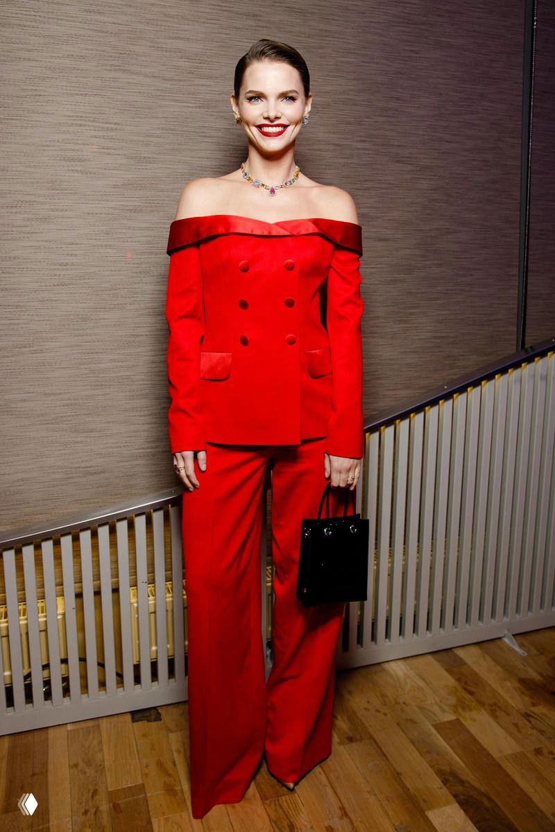 Женщина в ярком красном брючном костюме позирует в гостиной обстановке: стильное платье‑костюм, яркая помада и элегантная стойка.