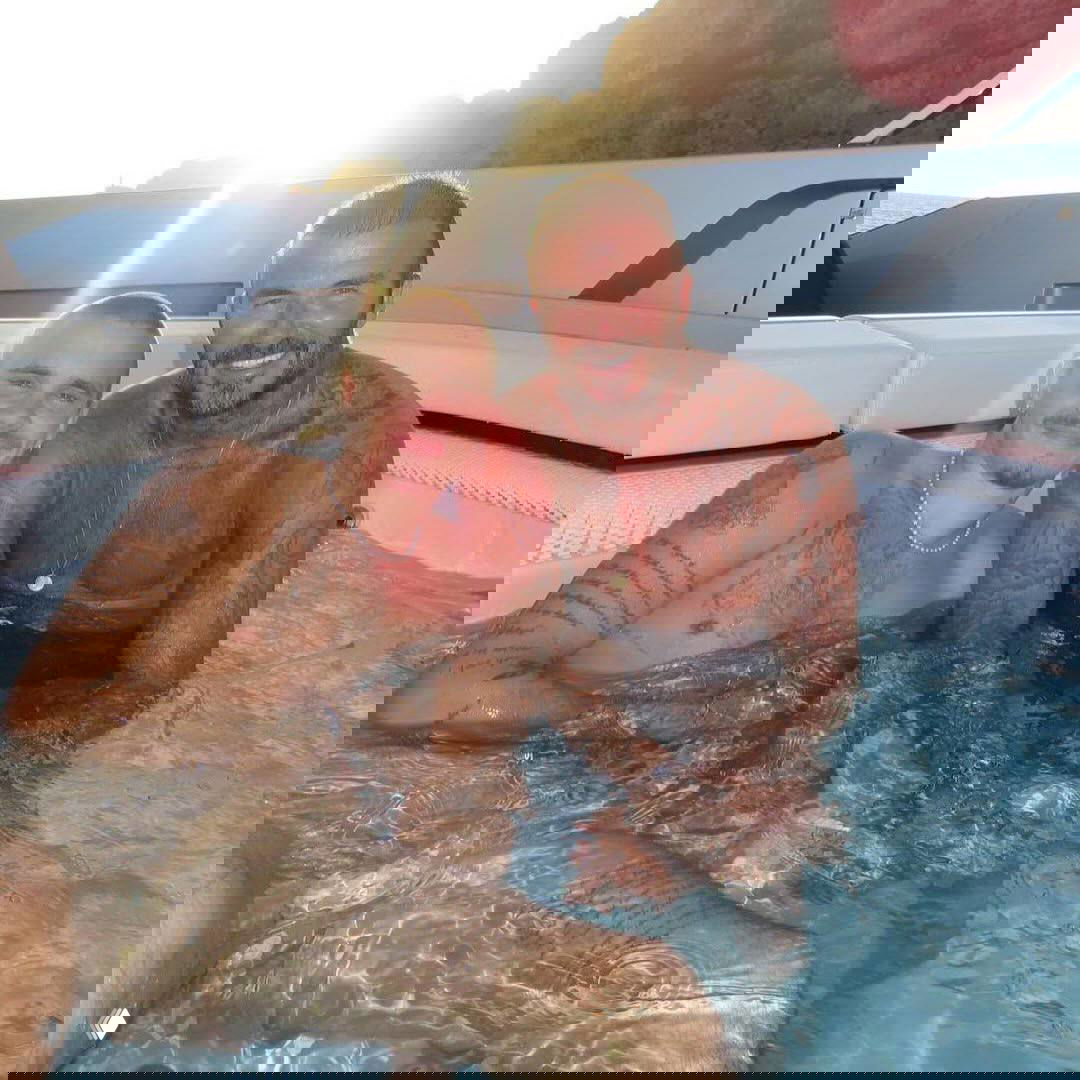 Двое мужчин в гидромассажной ванне на яхте, один с заметными татуировками, фон — морской берег и заходящее солнце.