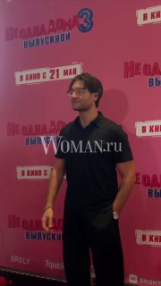 Фото DAVA в чёрной футболке на красном фоне фотозоны премьеры фильма «Не одна дома 3. Выпускной», заметен логотип Woman.ru и рекламный баннер