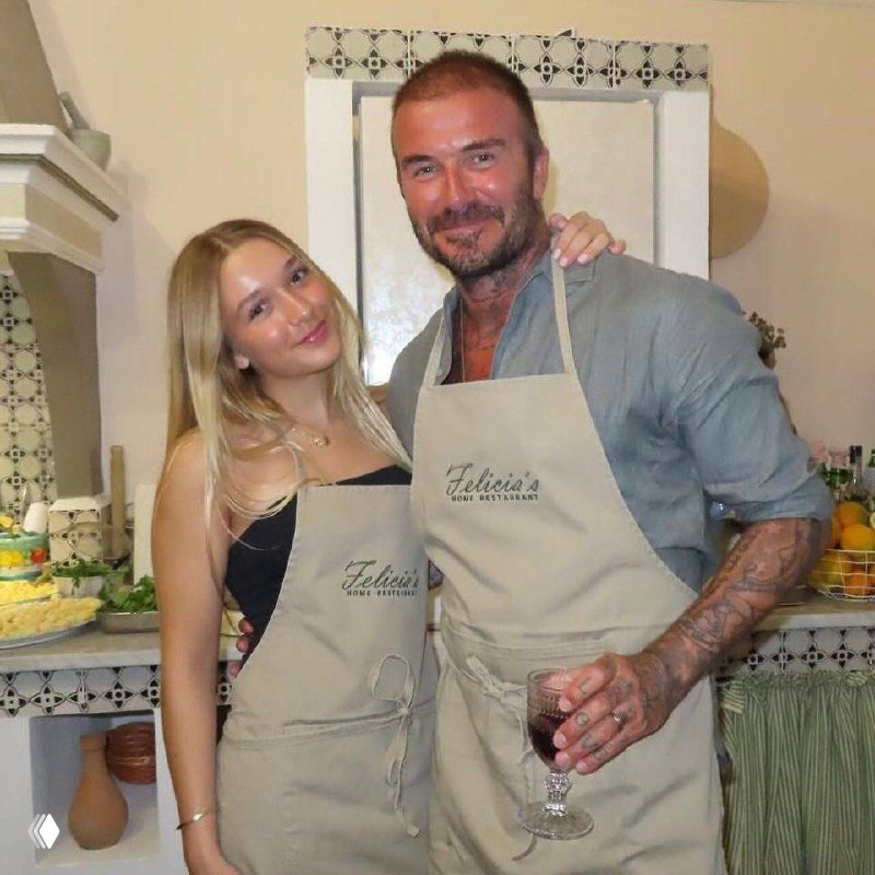 Мужчина улыбается на кухне рядом с молодой женщиной; оба в фартуках на фоне домашней сервировки и продуктов.