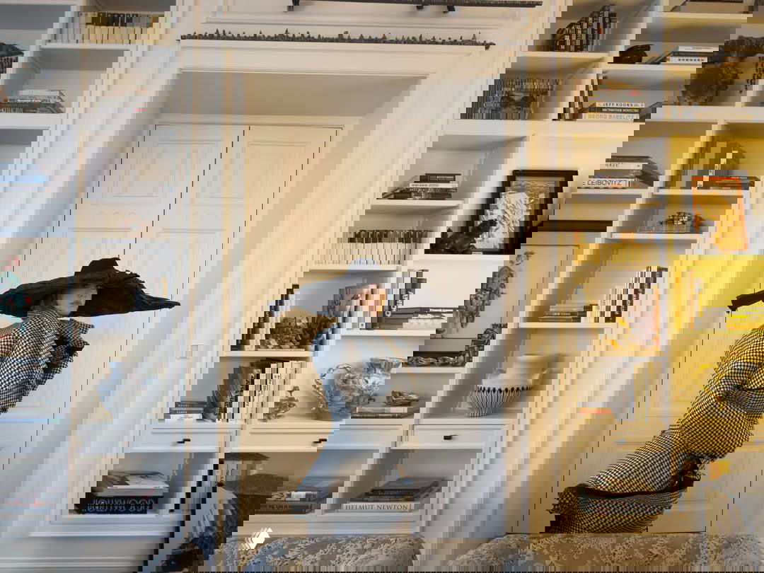 Вертикальный кадр у дверного проёма: высокий стеллаж с книгами и антиквариатом, на переднем плане тканевые кресла и декоративные предметы интерьера