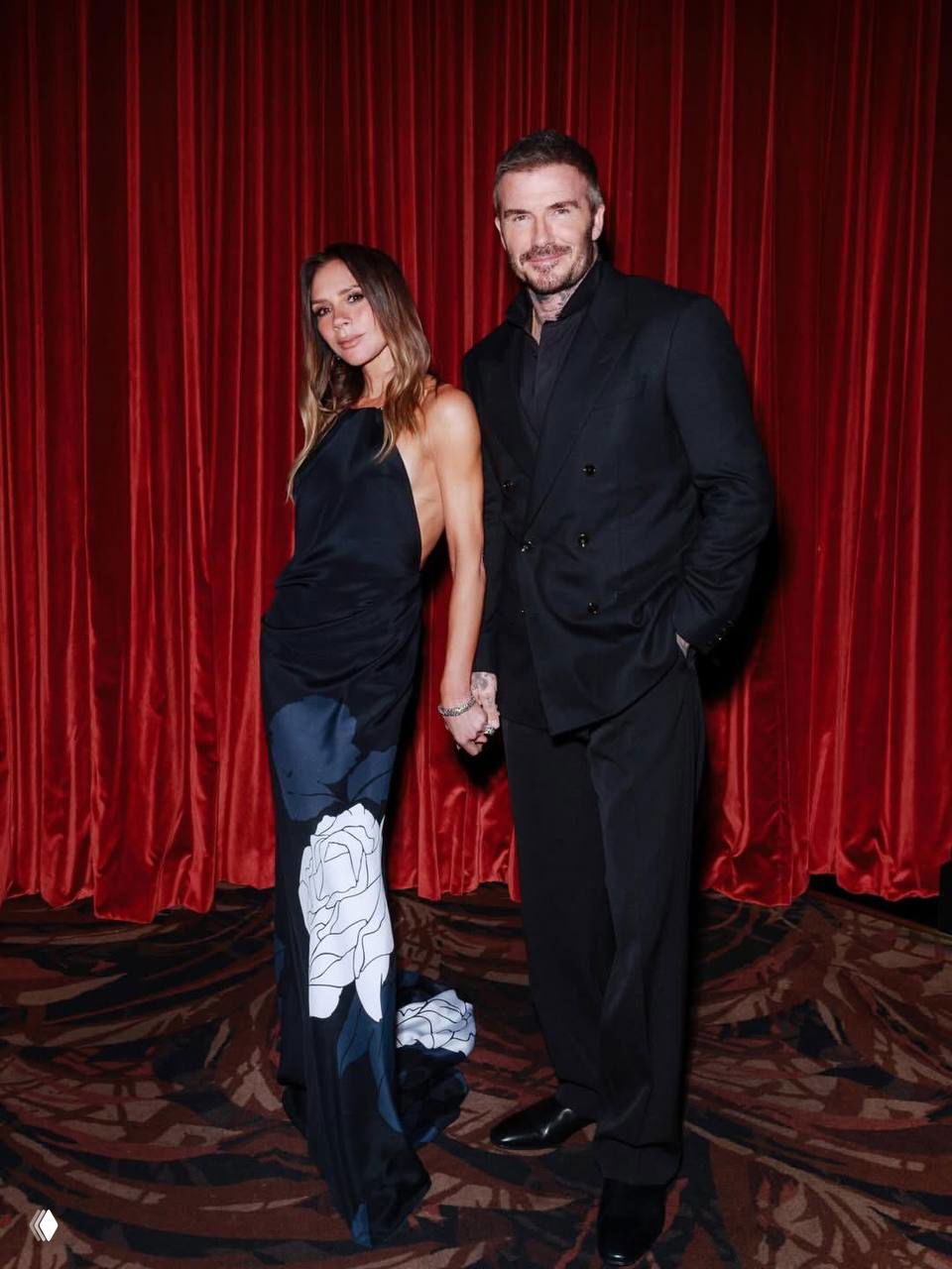 Пара держится за руки в элегантных вечерних образах на фоне красной бархатной занавески, официальный снимок во время торжественной вечеринки.