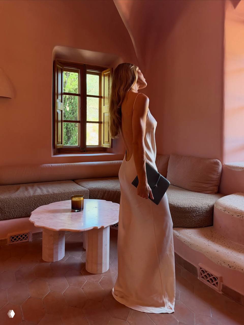 Модель в длинном бежевом платье, вид со спины в интерьерном кадре: розовая стена, окно с видом на сад, элегантная поза и мягкий свет.
