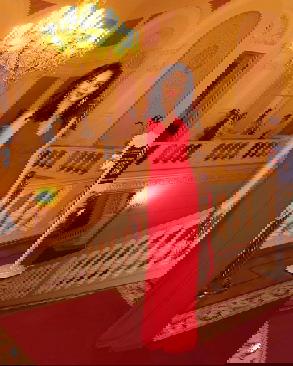 Женщина в длинном красном платье стоит у парадной лестницы Большого театра в украшенном праздничными гирляндами интерьере.