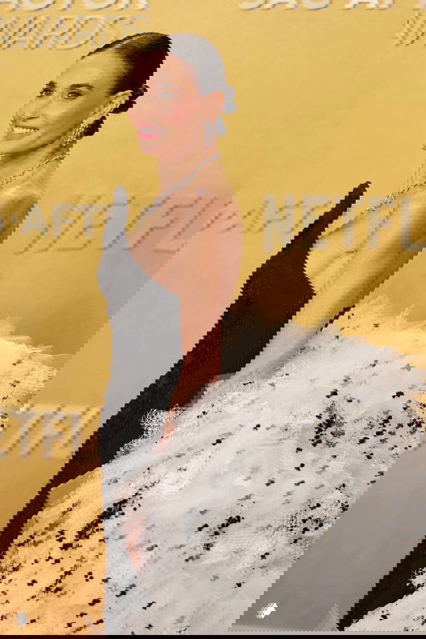 Профиль женщины в черном платье и дорогими украшениями, пышная белая тюлевая деталь и логотипы спонсоров на фоне.