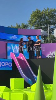 На летней сцене фестиваля стоят люди у яркой инсталляции с логотипом Woman.ru; видна цветная детская горка.