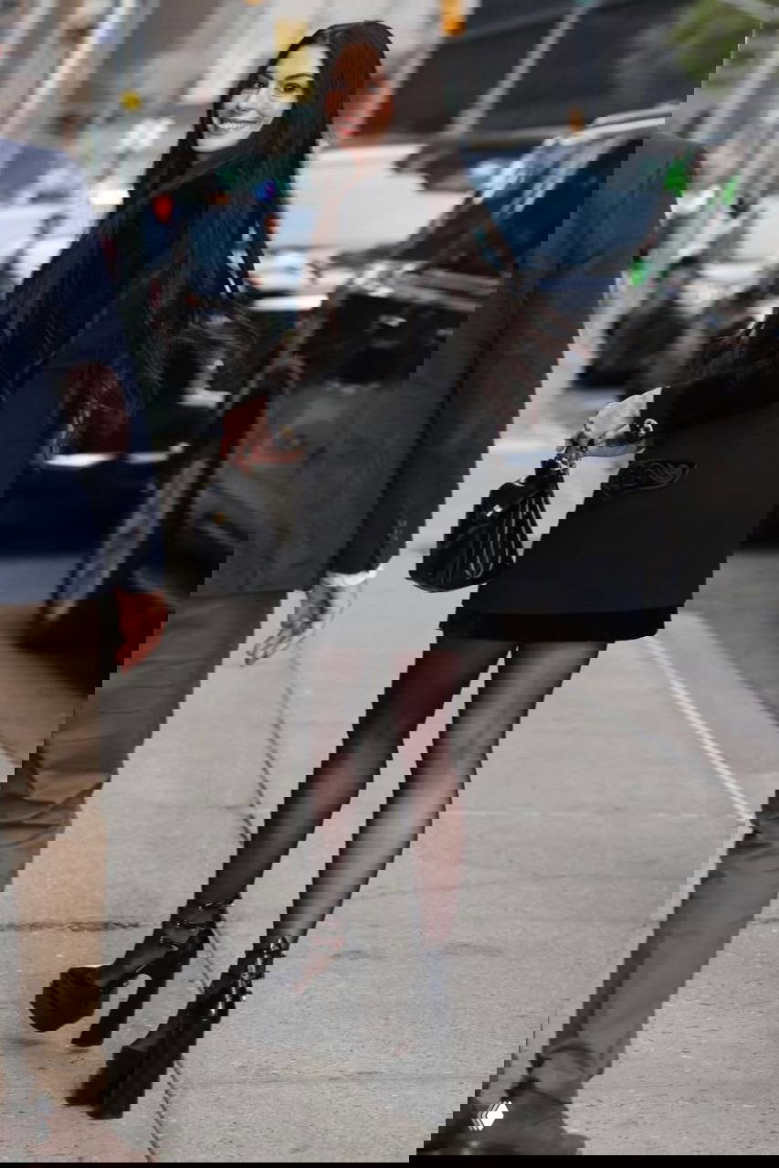 Кадр сзади: Энн Хэтэуэй в чёрном мини‑пальто и чулках уходит по улице между припаркованными автомобилями и прохожими, видна длина волос и силуэт.