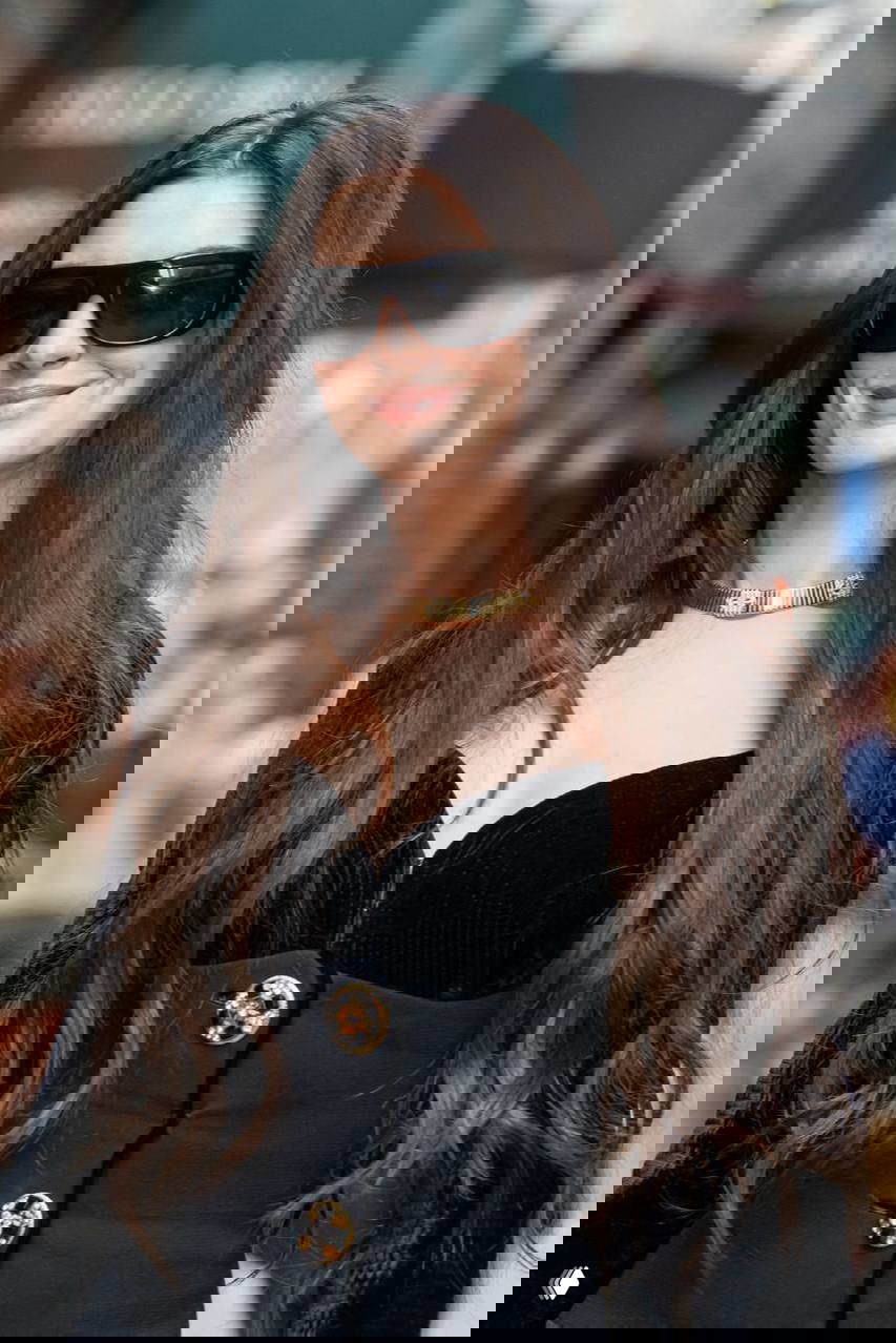 Крупный план Энн Хэтэуэй в чёрном корсетном топе с золотым колье и длинными тёмными волосами на фоне городского пейзажа и прохожих.