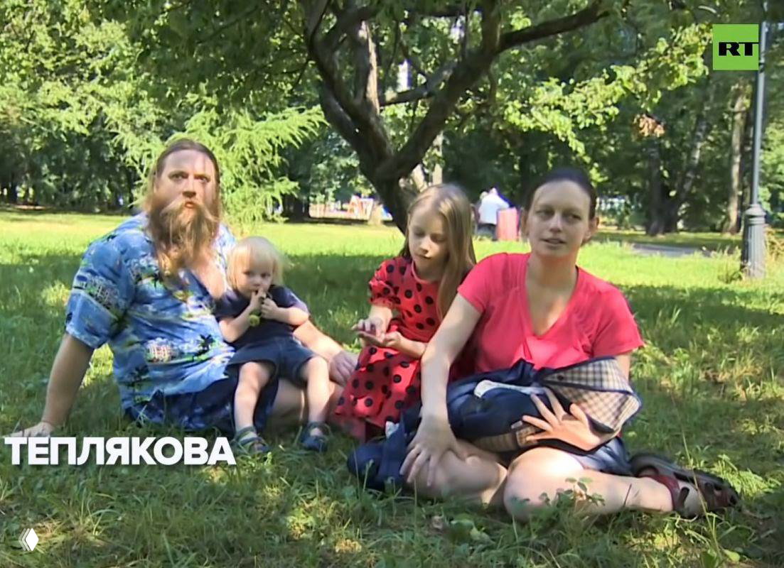 Семейная сцена в городском парке: мужчина, женщина и двое детей сидят на траве летом, видно зелёную листву и логотип вещателя в углу кадра.