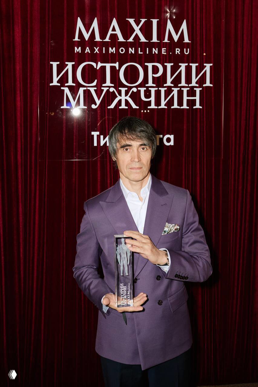 Портретный кадр лауреата в фиолетовом пиджаке, держащего хрустальную статуэтку на фоне фотостены проекта «Истории мужчин»