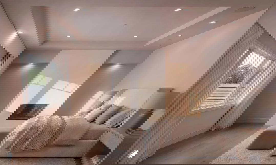 Просторная светлая спальня особняка: большая кровать, камин в нише, мягкие текстильные элементы и естественный дневной свет через высокие окна.