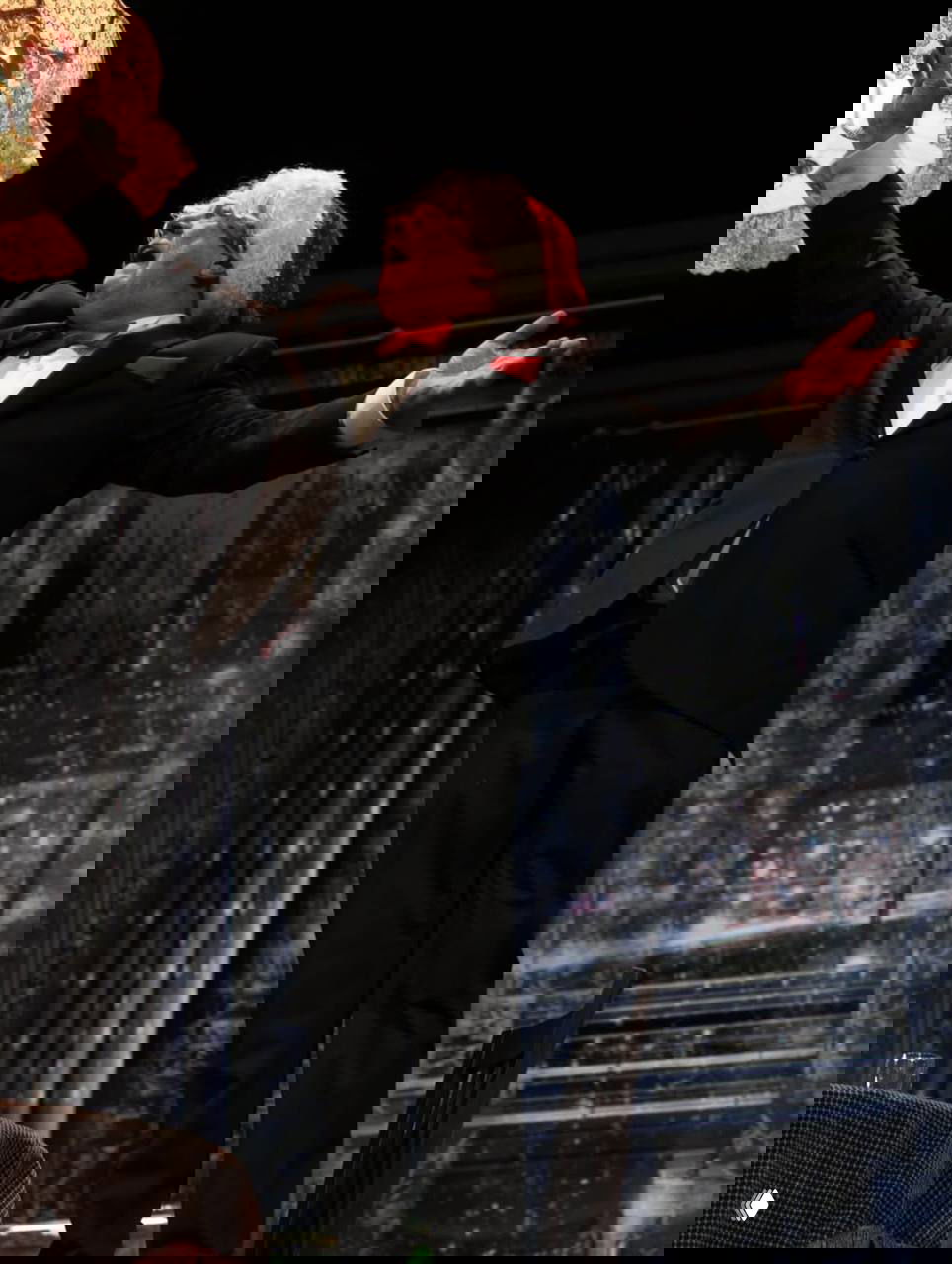 Актёр в смокинге и рождественской шапке, раскинув руки на сцене театра во время эмоционального эпизода спектакля