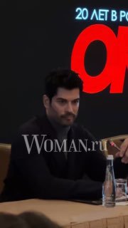 Крупный план Бурака Озчивита на пресс‑конференции в Москве: лицо актёра, бутылка воды на столе и тёмный фон с логотипом, заметен ватермарк Woman.ru.