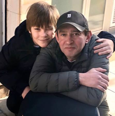 На фото мужчина в тёмной куртке и бейсболке обнимает мальчика; кадр близкий, передаёт домашнюю, тёплую атмосферу на фоне фасада здания.