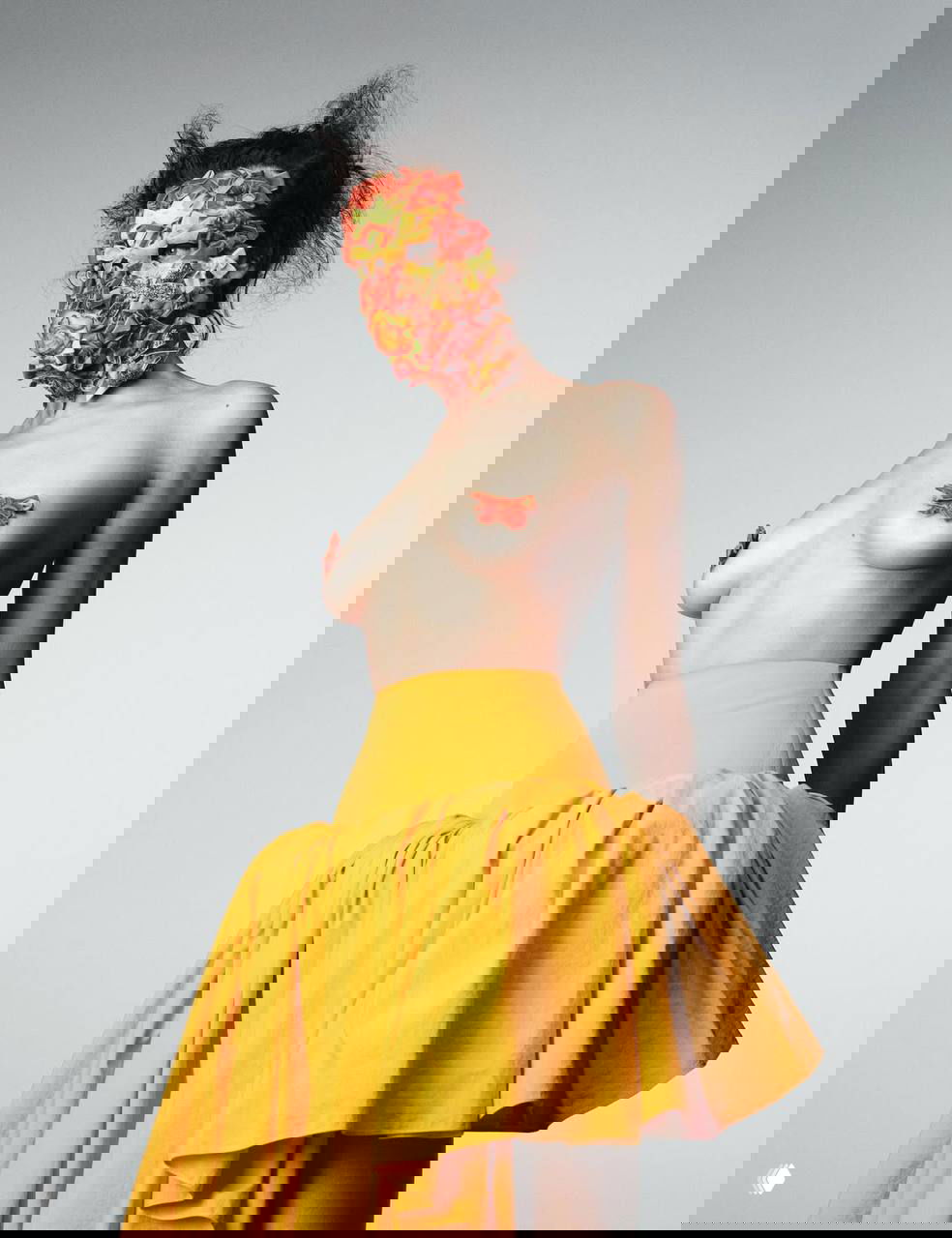 Фотография Ирины Шейк в жёлтой юбке и маске из цветочных элементов: обнажённый торс частично декорирован, снимок стилизован в авангардной эстетике журнала.