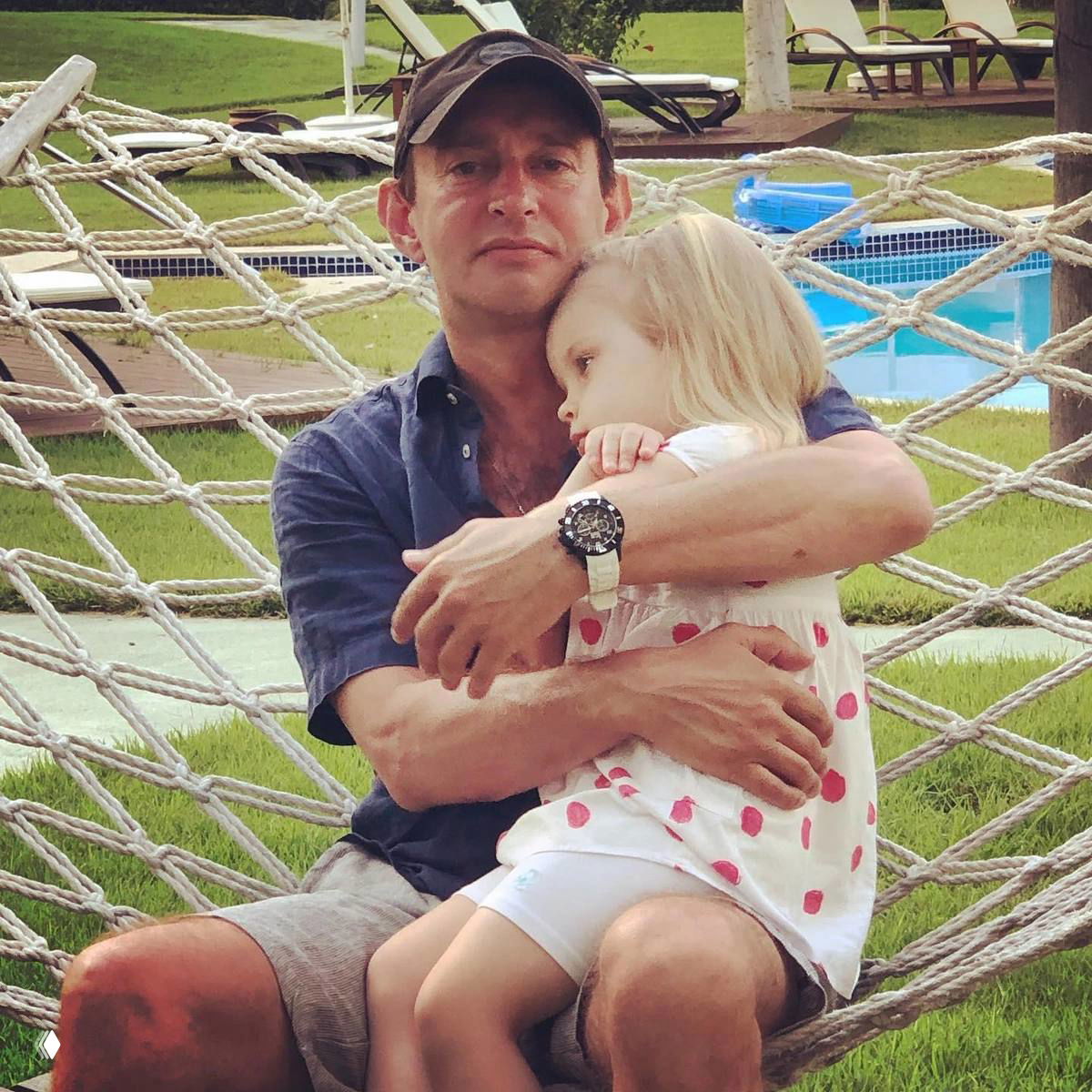 На фото мужчина в кепке сидит в гамаке у бассейна и держит на руках маленькую девочку в лёгком платье; видна зелёная лужайка и шезлонги.