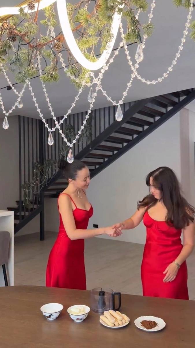 Две женщины в красных платьях снимаются в светлом просторном помещении особняка: видна лестница, большая люстра и стол с посудой и закусками.