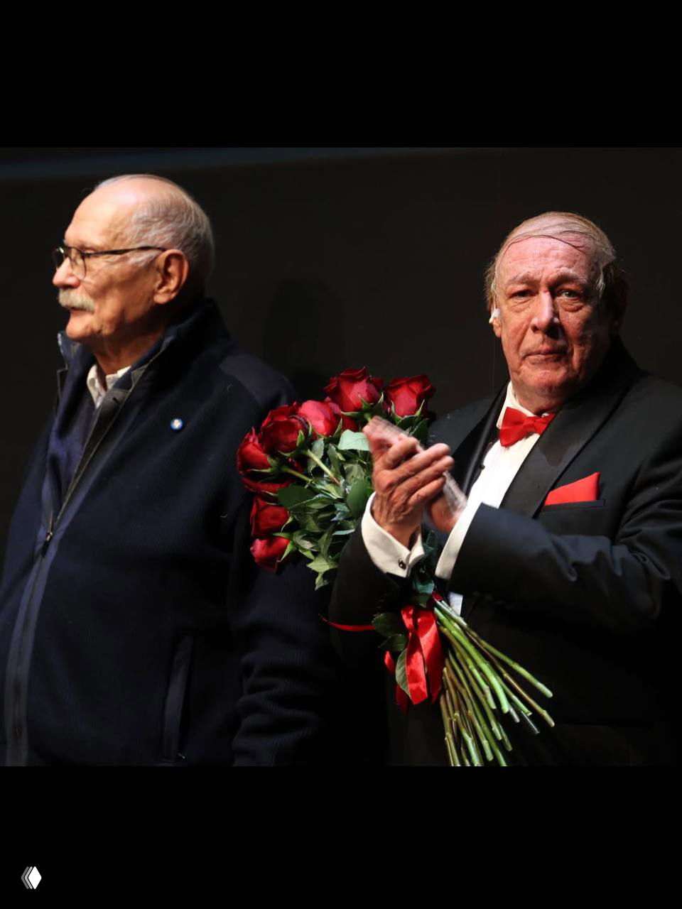 После спектакля: мужчина в смокинге с букетом красных роз аплодирует на сцене; рядом другой пожилой мужчина.