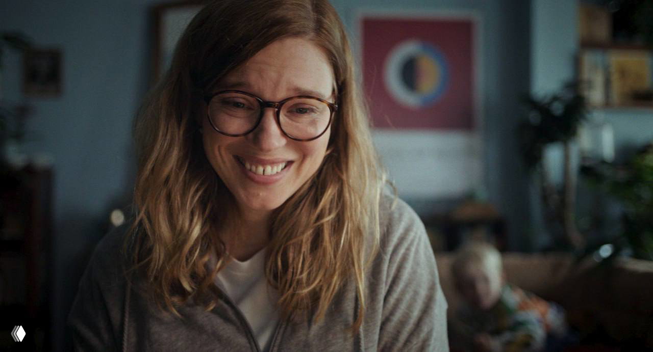 Кадр из фильма: улыбающаяся женщина в очках в домашней обстановке, тёплая интерьерная сцена, подчёркнутая естественной кинематографией.