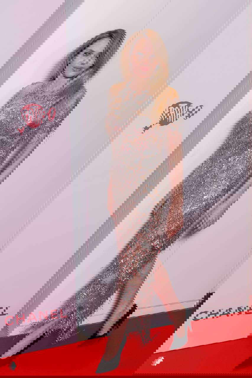 Полноразмерный кадр: женщина в длинном блестящем платье идёт по красной дорожке на фоне рекламных панелей и логотипов кинособытия в Париже.