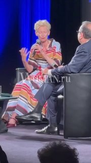 Женщина в полосатом платье сидит на сцене фестиваля и говорит в микрофон на фоне синей подсветки, кадр снят из зала