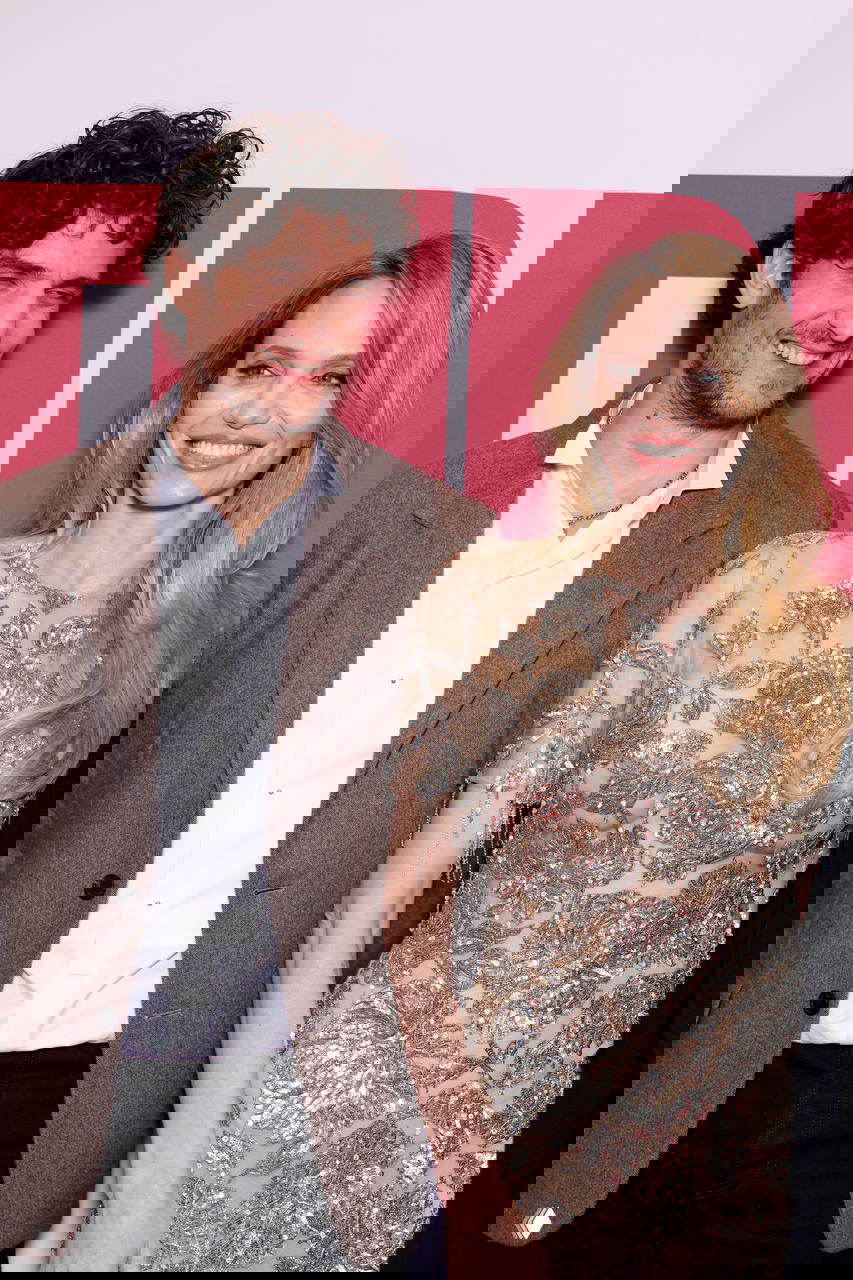 Женщина улыбается рядом с мужчиной на красной дорожке; видно декоративное серебристое платье и атмосферу фотоколла мероприятия.