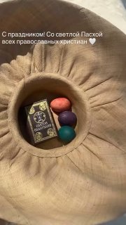 Скриншот из сторис: шляпа с тремя крашеными яйцами и миниатюрной иконкой внутри, подпись с поздравлением к Пасхе на фоне ткани.