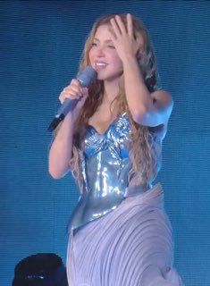 Крупный план Шакиры в сценическом образе с микрофоном и развивающимися волосами: момент выступления и эмоции певицы на концерте.