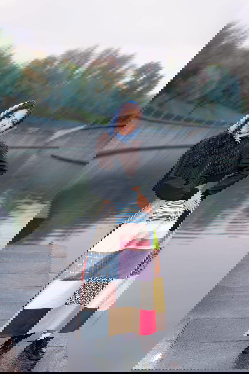 Модель в пэчворк-юбке на набережной у спокойного канала; осенний пейзаж и отражение воды на заднем плане.