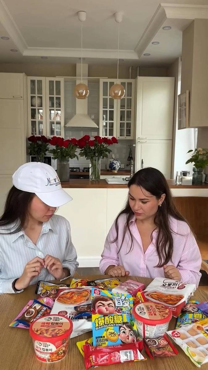 Две женщины сидят за кухонным столом особняка, перед ними набор разноцветных снэков и упаковок, на заднем плане светлая кухня и букет роз.