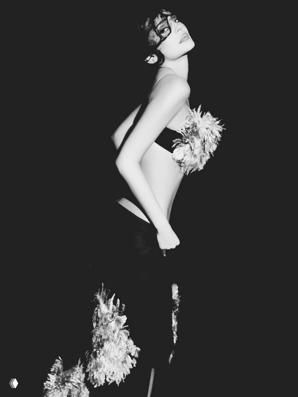 Чёрно‑белый кадр: модель в эффектной позе с декоративными перьями на талии на тёмном фоне, стилизованная студийная фотография.