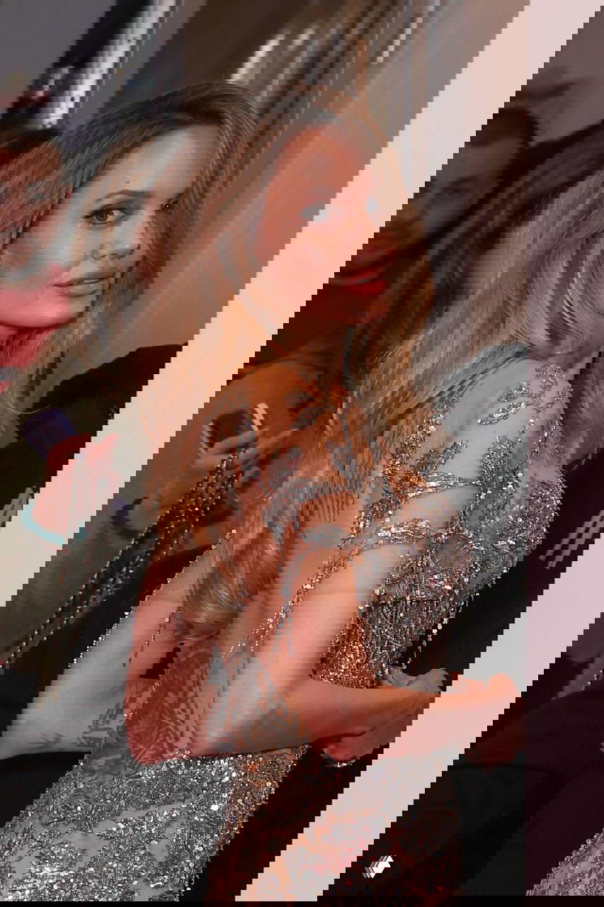 Репортёрский кадр в коридоре: женщина в расшитом платье показывает татуировку на спине и уходит за кулисы после премьеры фильма.