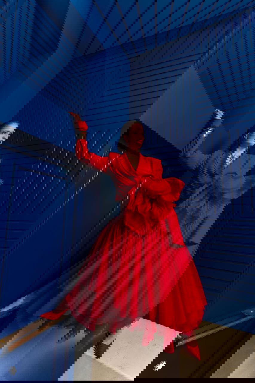 Женщина в ярком красном платье и туфлях позирует на фоне строгой синей архитектуры — контрастный студийный кадр для фотосессии.