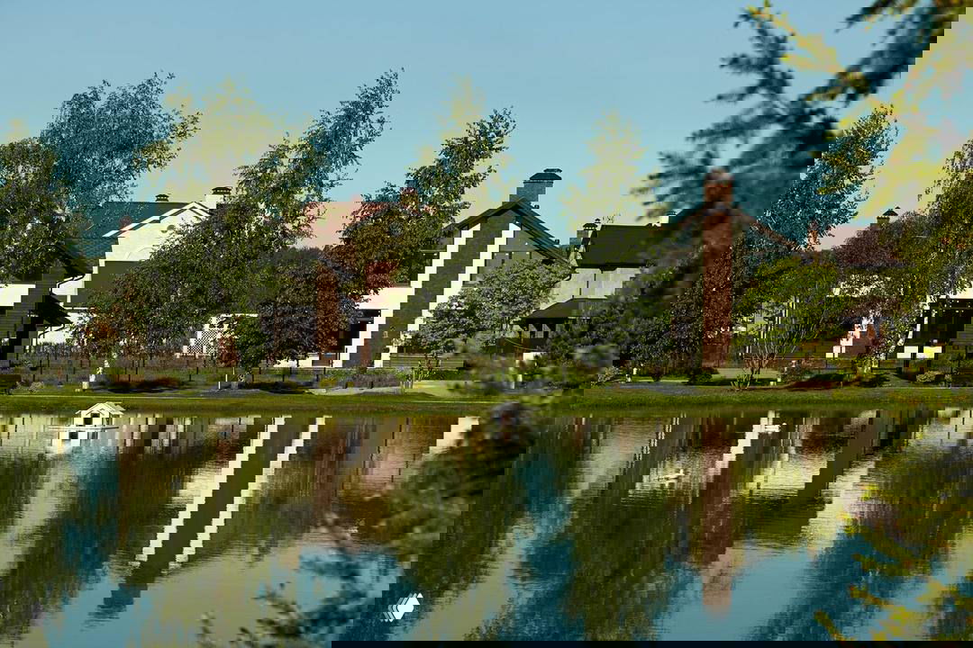 Широкий вид на берег озера и несколько коттеджей в Новорижском поселке, дом на заднем плане и спокойная водная гладь с утками.