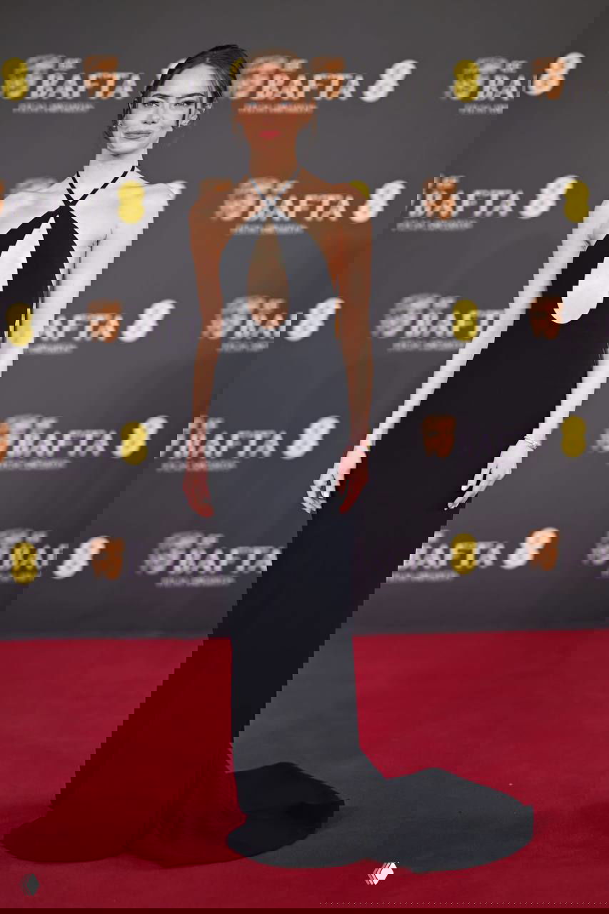 Молодая женщина в облегающем чёрном платье с глубоким вырезом позирует на красном ковре BAFTA на фоне логотипов премии.