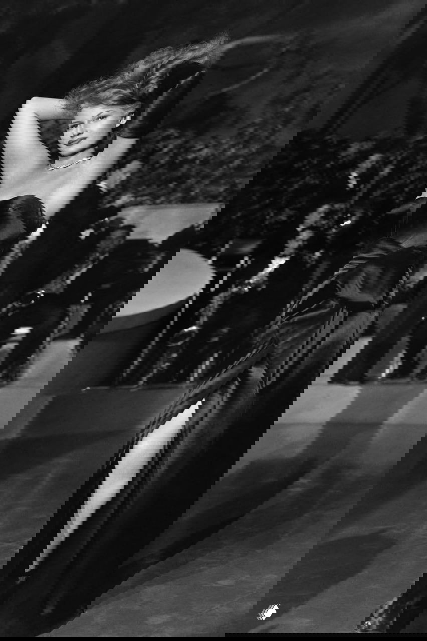 Чёрно‑белый кадр: актриса в атласном платье с высоким разрезом позирует на танцполе, позади виден оркестр.