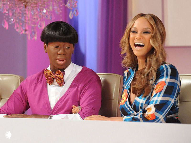 Джей Александр и Тайра Бэнкс сидят рядом за столом в студии, обе улыбаются; фрагмент из телепроекта, связанного с модельным шоу.