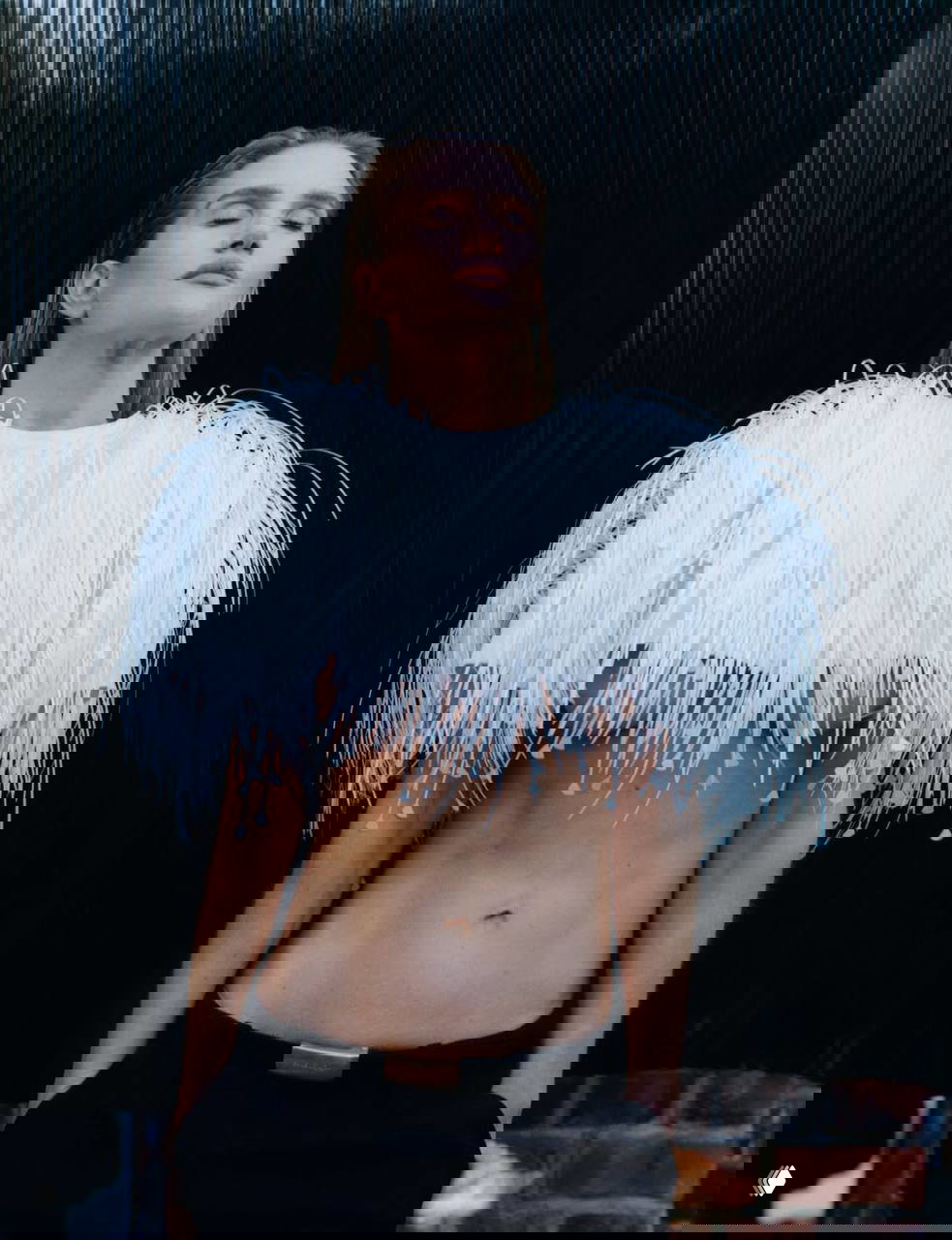 Модель в коротком топе с декоративными перьями и открытым животом позирует на тёмном фоне — показаны формы и текстура одежды.