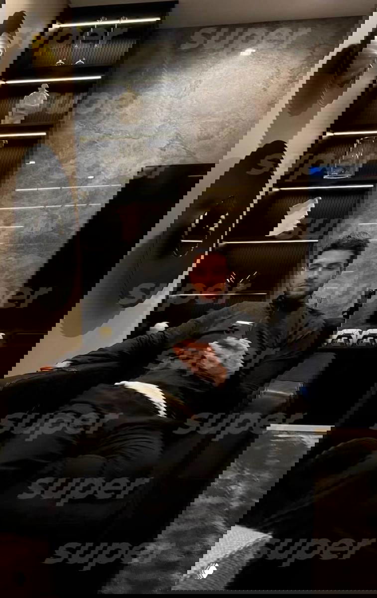 Мужчина сидит в кресле в интерьере с мраморной стеной и полками; держит смартфон, в кадре заметен водяной знак «super», роскошная обстановка.