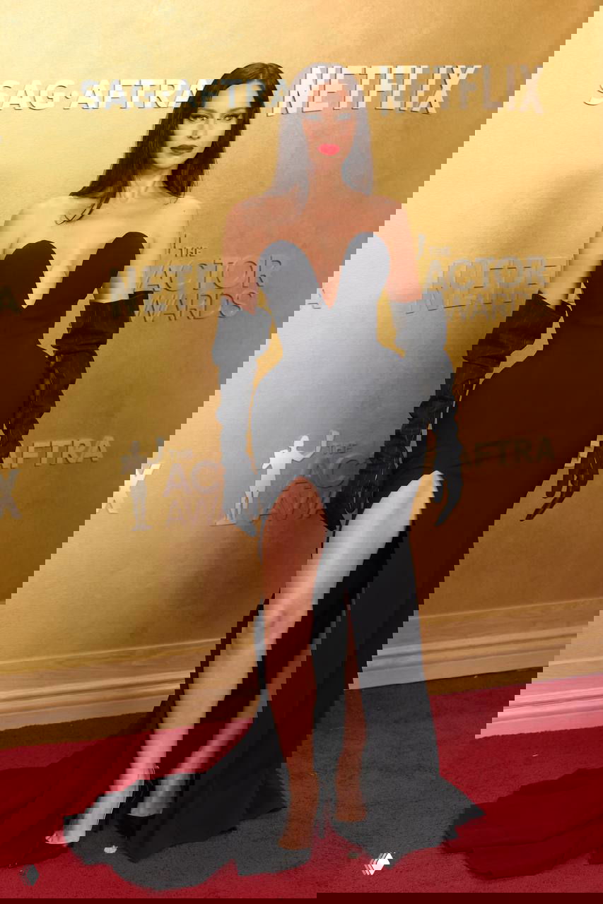 Фотография в полный рост: чёрное платье с высоким разрезом и длинными перчатками, туфли на каблуке — поза на красном ковре у логотипов мероприятия.