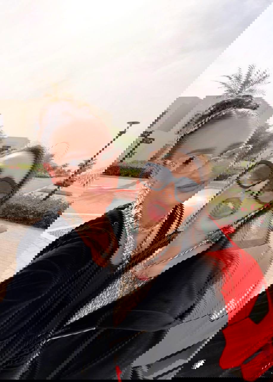 Селфи двух женщин на улице у красного спортивного автомобиля; обе в солнцезащитных очках и с крупными украшениями, улыбаются на фоне жилой застройки.