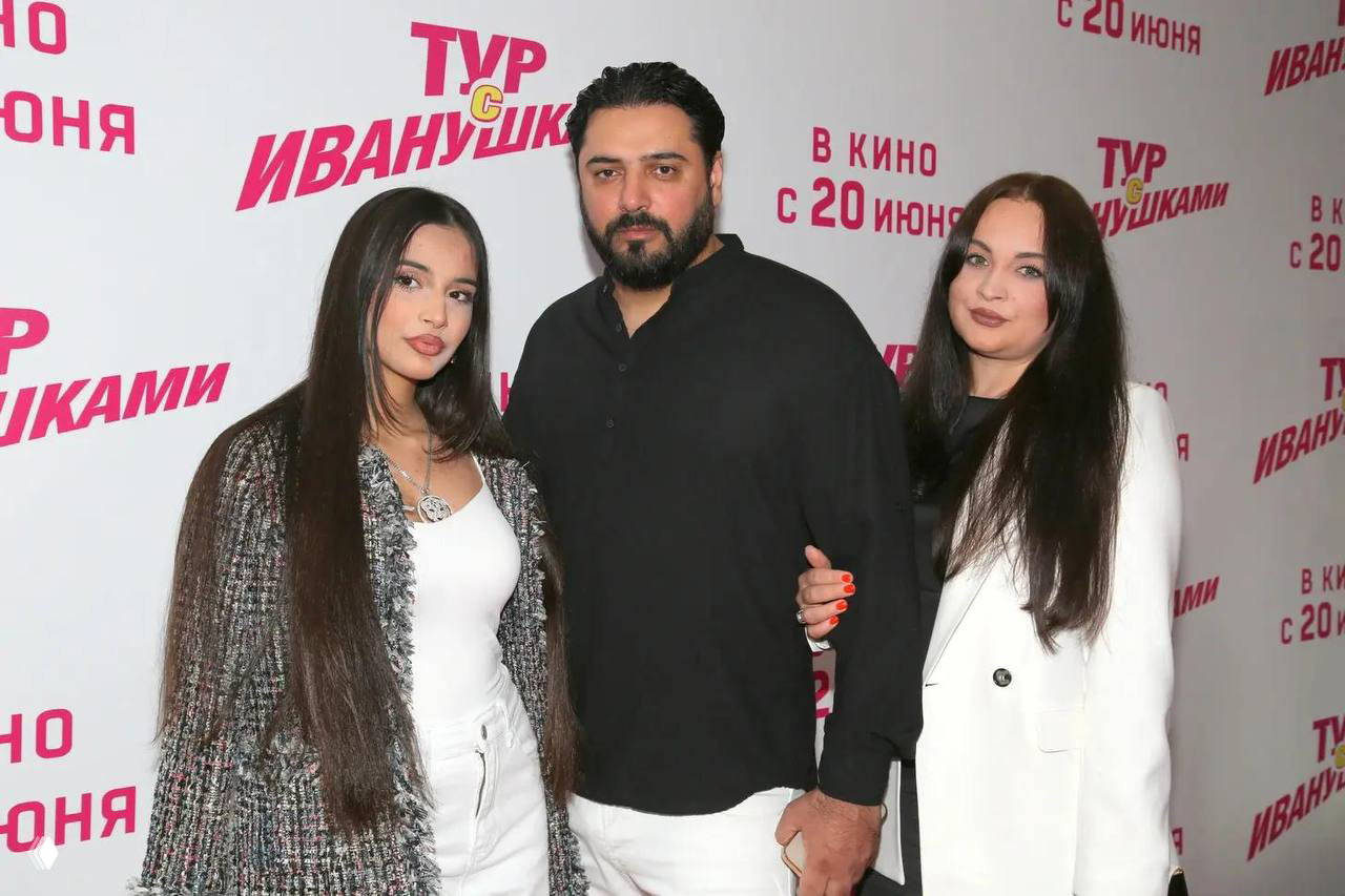 Премиальное групповое фото: мужчина и две женщины позируют на фоне рекламного баннера кинопоказа, вечерняя обстановка и праздничные улыбки.