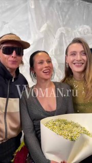 Катя IOWA улыбается рядом с двумя коллегами на фоне съёмочной площадки; видно букет и атмосферу дружеской встречи в шоу-бизнесе.