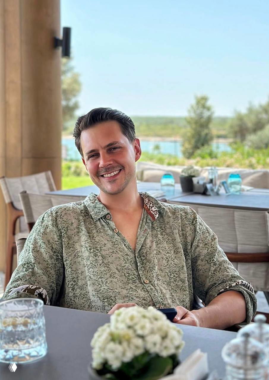 Улыбающийся Милош Бикович за столиком на террасе у воды, летняя атмосфера, сервировка и природный фон на заднем плане.