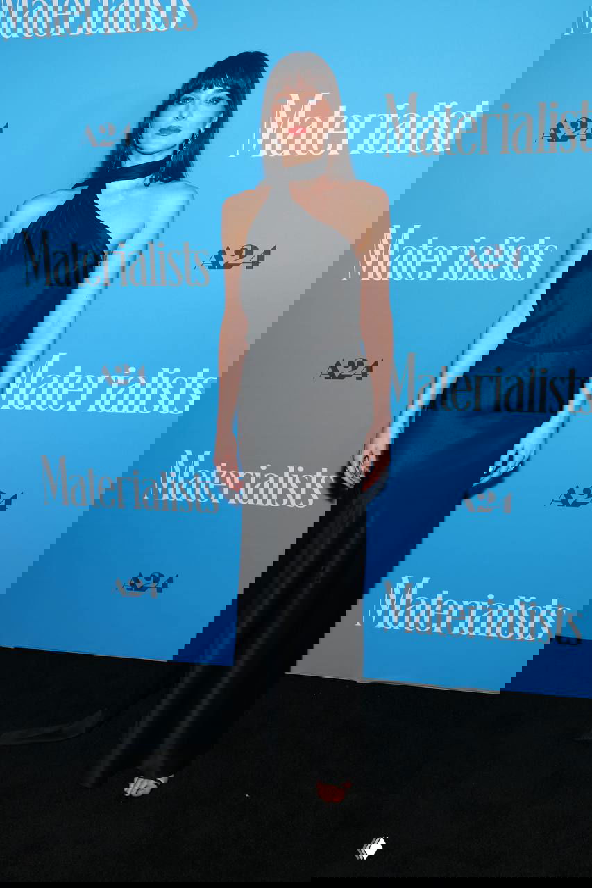 Женщина в чёрном атласном платье с асимметричным вырезом на одно плечо; минималистичный вечерний образ на фоне пресс-волла мероприятия.