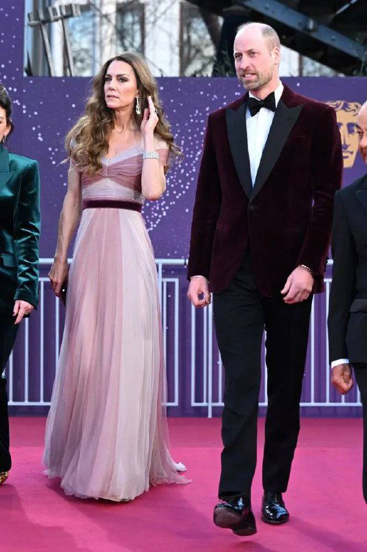 Полный рост женщины в воздушном розовом платье и мужчины в тёмном смокинге на красной дорожке BAFTA — общий план их появления.