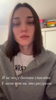 Вертикальное видео/сторис: женщина в серой толстовке смотрит в камеру; видна полка с бытовыми предметами и нейтральный домашний фон.